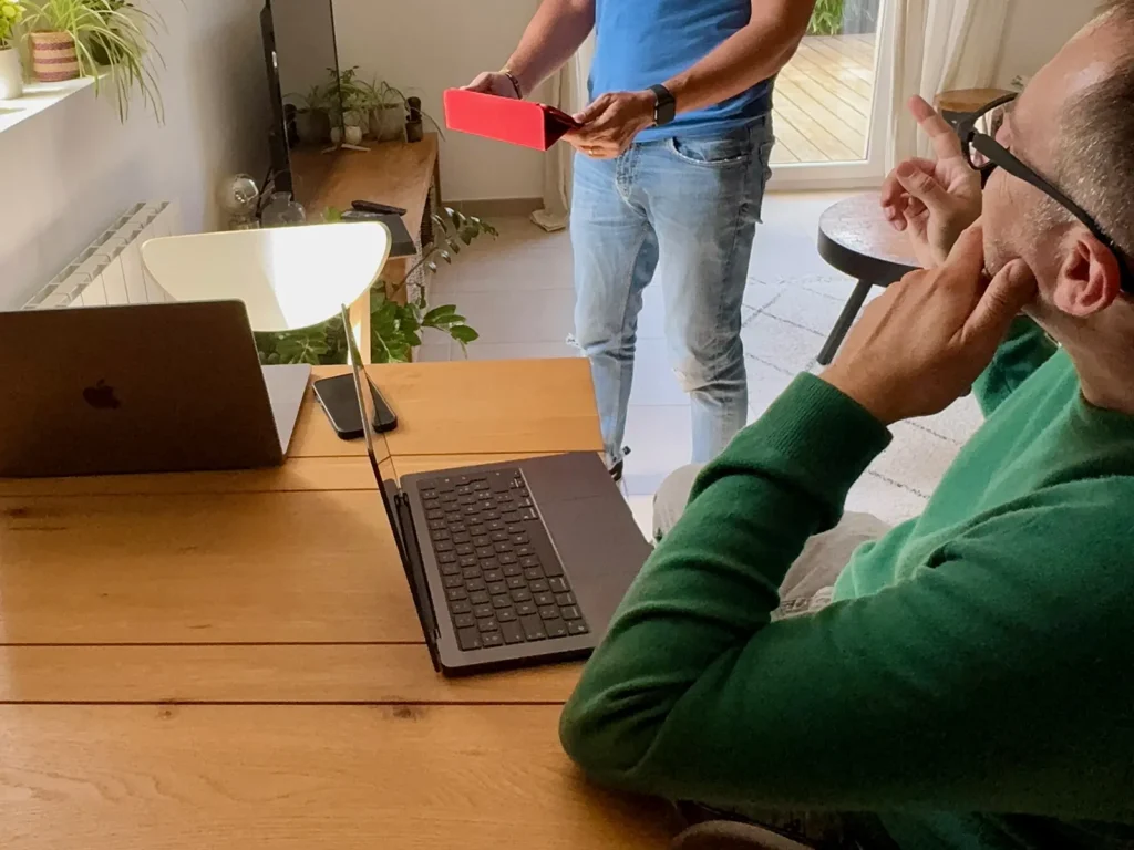 DAMIEN IBELIEVE 10 Deux personnes discutent dans un salon moderne, avec des ordinateurs portables et une tablette rouge, entourés de plantes et de meubles clairs.