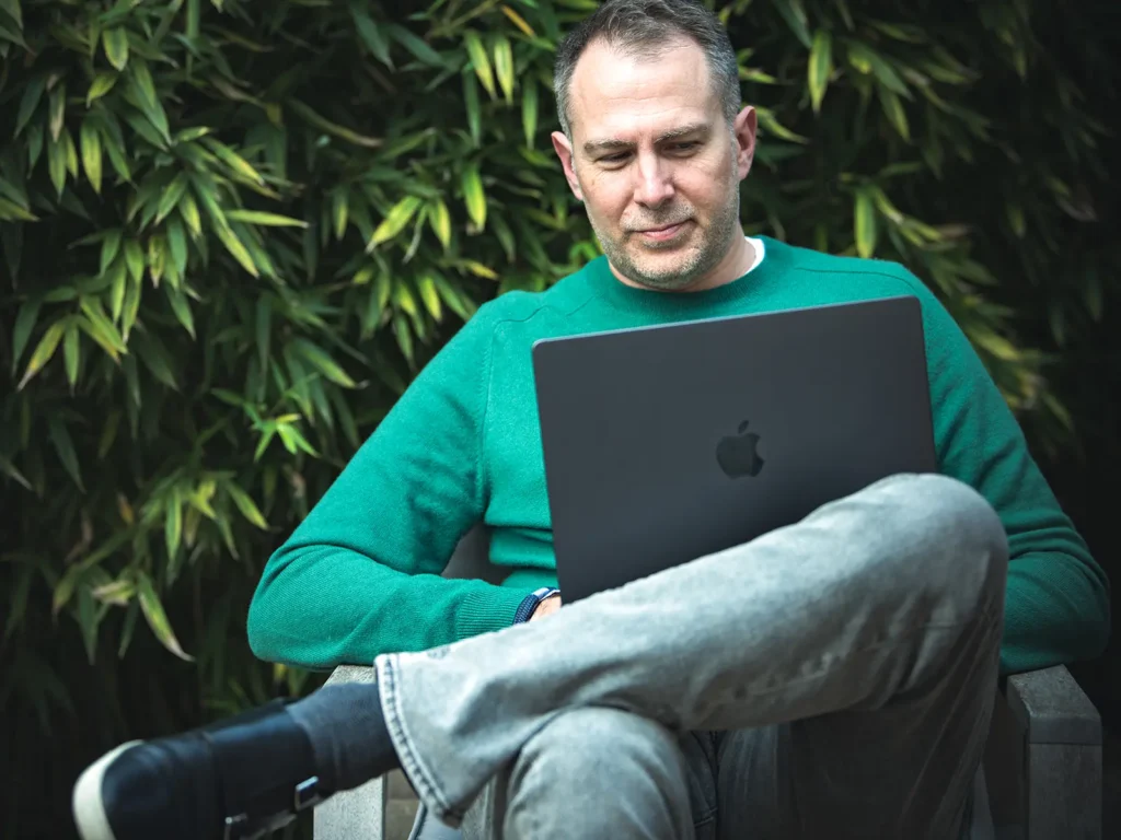 Damien formateur de chez IBELIEVE est assis et regarde un ordinateur portable devant une haie verte.