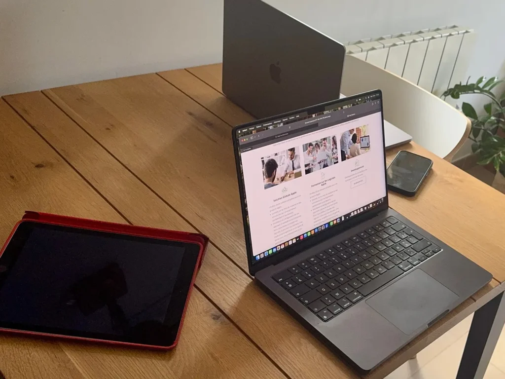 DAMIEN IBELIEVE 8 Trois ordinateurs portables et une tablette sur une table en bois, près d'un radiateur et d'une plante, dans un espace de travail.