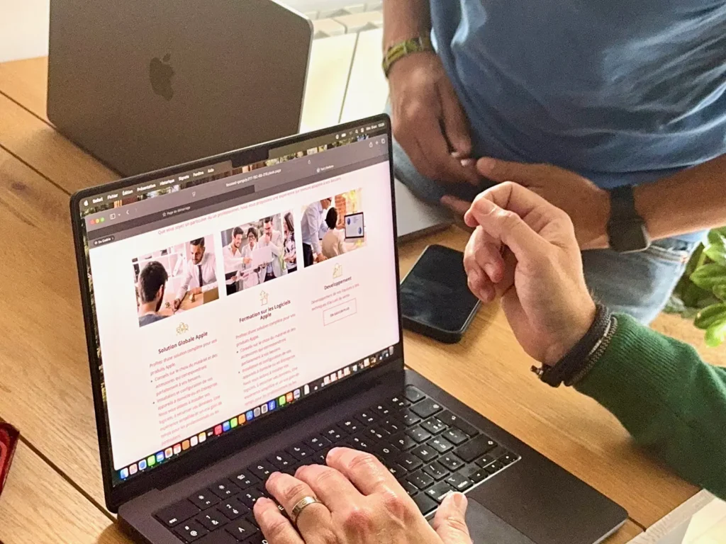 DAMIEN IBELIEVE 9 Deux personnes discutent devant un ordinateur portable affichant un site web. Un smartphone repose sur la table en bois à côté.